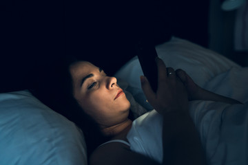 Insomnia and social media addiction concept. Young woman uses smartphone while lying in bed at night
