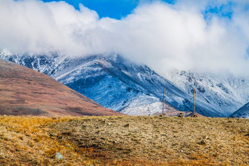 Russia. Altai. Yellow grass and stones in the foreground, in the background mountains in the clouds covered with snow.