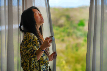 lifestyle portrait of young beautiful happy and relaxed Asian Korean woman in stylish bathrobe by luxury hotel room window having morning coffee enjoying the tropical view