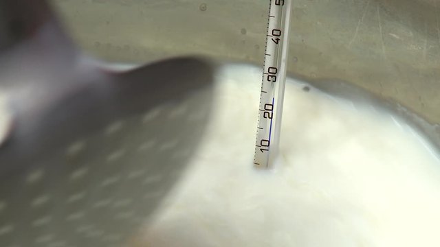 Handheld, close up shot of a thermometer in milk as a large metal spoon stirs.