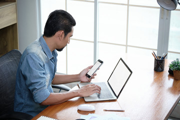 Bearded man sitting at a desk in his house looking at the cellphone and laptop. Online business entrepreneur checking the order list on the internet platform while light shining through the window.
