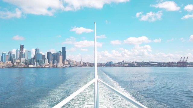 Steady view of Downtown Seattle, CenturyLink field, T-Mobile Park, and Port of Seattle from water off stern of Washington State Ferry.  Wake is seen behind steering pole on sunny summer day.