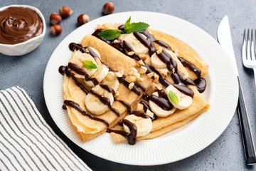 French style crepes with banana, chocolate sauce and chopped nuts on a white plate. Tasty sweet dessert