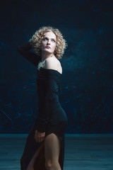 Fototapeta premium Young male blond androgyne in the image of a beautiful woman posing in a black dress on a dark background 