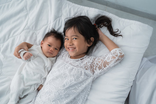 Asian Daughter Kid With Infant Sibling Playing On Bed Wearing White Together