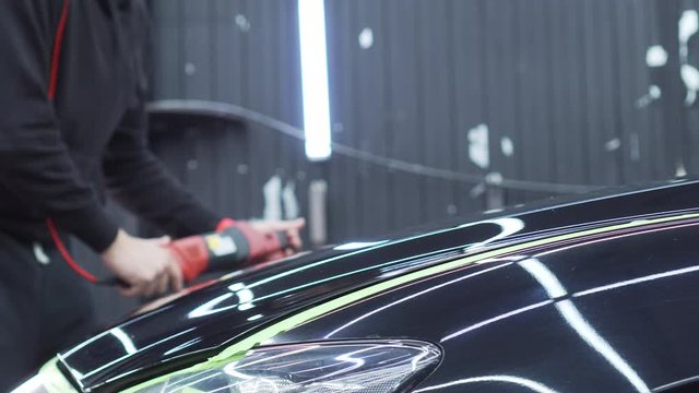 A male polisher works on polishing a black car in a well-lit but shabby garage