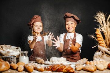Adorable girl with brother in chief hats and aprons cooking at table with bread loaves making fresh dough and having fun