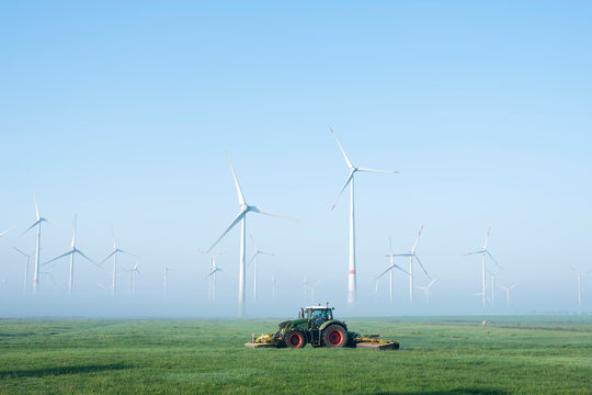 farmer mows grass near wind turbine farm in oastfriesland on misty summer morning
