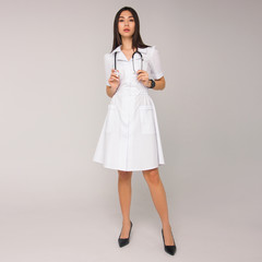 young stylish brunette nurse with long hair in white medical dress with stethoscope on her neck is standing on the white wall background. medical fashion concept. free space