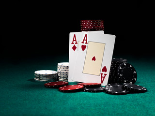 Two aces hearts and diamonds standing leaning on chips piles, some of them laying nearby on green cover of playing table. Black background. Close-up.