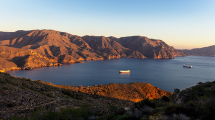 The morning sun shines into the bay at Cabo Tinoso near the Spanish port city of Cartagena. Large ships moored in the bay between the mountains.