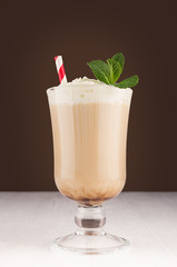 Macchiato coffee with layers, cream, red striped straw and green fresh mint in transparent goblet on dark brown background and white wooden table.