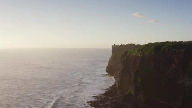 Flying sideways, aerial view of picturesque high cliffs above blue ocean. Foamy waves. Silhouette of Uluwatu temple on the cliff top. Bali, Indonesia