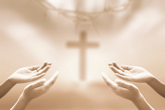 Human hands reaching out in prayer towards a blurred Christian cross and crown of thorns