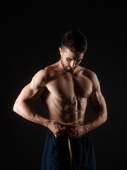 Fototapeta na wymiar Handsome muscular man with measuring tape on dark background. Weight loss concept