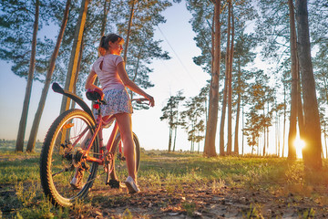 young girl with a bicycle on an autumn pine forest. sunset sun