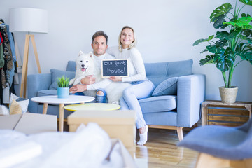 Young beautiful couple with dog sitting on the sofa holding blackboard with message at new home around cardboard boxes