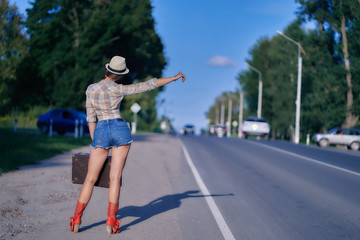 young hipster woman in shorts and red boots is waiting for a ride on the road. hitchhiking around the country.