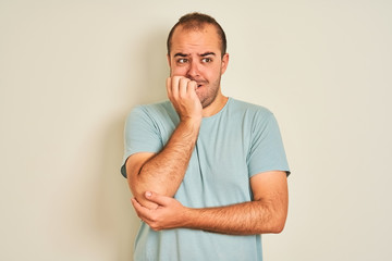 Young man wearing blue casual t-shirt standing over isolated white background looking stressed and nervous with hands on mouth biting nails. Anxiety problem.