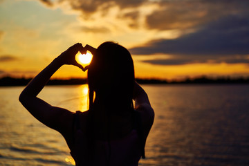 silhouette of a girl. in the setting sun on the lake. hands depicts a heart shape.