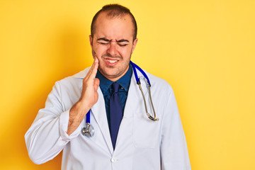 Young doctor man wearing coat and stethoscope standing over isolated yellow background touching mouth with hand with painful expression because of toothache or dental illness on teeth