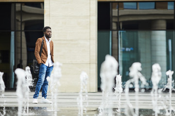 Full length portrait of pensive African-American businessman standing outdoors by fountain in city...