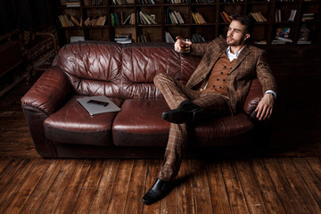 young successful man in a brown kosmtyum drinks whiskey. bearded businessman sitting in a library...