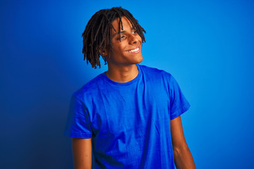 Fototapeta na wymiar Afro american man with dreadlocks wearing t-shirt standing over isolated blue background looking away to side with smile on face, natural expression. Laughing confident.