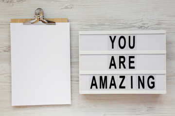 'You are amazing' words on a modern board, clipboard with blank sheet of paper on a white wooden...