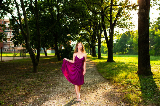 Beautiful curly hair young woman wearing elegant purple dress standing on a path in the middle of forest with rays of sunlight during sunset