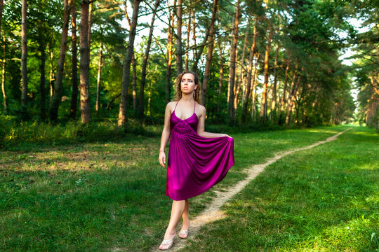 Beautiful curly hair young woman wearing elegant purple dress standing on a path in the middle of forest with rays of sunlight during sunset