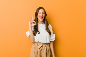 Young caucasian woman winks an eye and holds an okay gesture with hand.