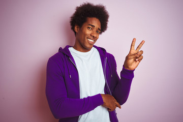 Young african american man wearing purple sweatshirt standing over isolated pink background smiling with happy face winking at the camera doing victory sign with fingers. Number two.