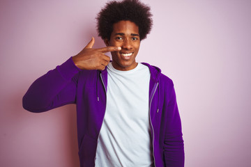 Fototapeta na wymiar Young african american man wearing purple sweatshirt standing over isolated pink background Pointing with hand finger to face and nose, smiling cheerful. Beauty concept