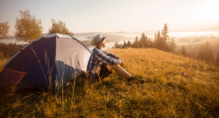 Hiker in hat sits near tent meets good morning on top of mountains