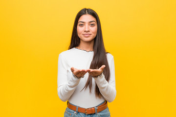 Young pretty arab woman against a yellow background holding something with palms, offering to camera.