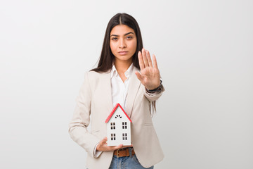 Young arab woman holding a house icon standing with outstretched hand showing stop sign, preventing...