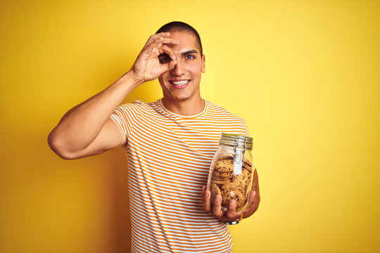 Young handsome man holding a jar of cookies over yellow isolated background with happy face smiling doing ok sign with hand on eye looking through fingers