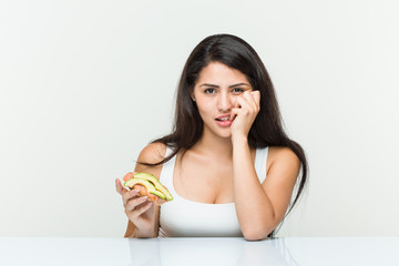 Young hispanic woman holding an avocado toast biting fingernails, nervous and very anxious.