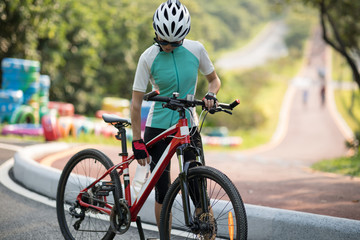Naklejka premium Riding on park bike path, woman cyclist drinking water while riding bike on sunny day