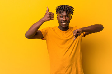 Young black man wearing rastas over yellow background showing thumbs up and thumbs down, difficult...