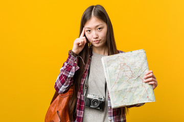 Young asian woman holding a map pointing his temple with finger, thinking, focused on a task.