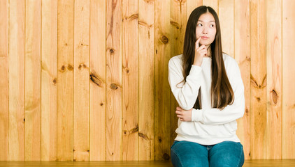 Fototapeta premium Young chinese woman sitting on a wooden place looking sideways with doubtful and skeptical expression.