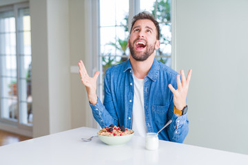 Handsome man eating cereals for breakfast at home crazy and mad shouting and yelling with aggressive expression and arms raised. Frustration concept.