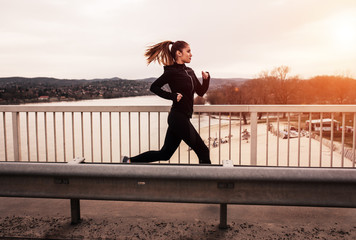 Young woman in black sports outfit running on the bridge in the city during sunset.