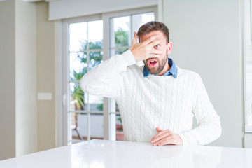 Fototapeta premium Handsome man wearing casual sweater peeking in shock covering face and eyes with hand, looking through fingers with embarrassed expression.