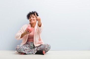 Young african american woman sitting with a piggy bank showing number one with finger.