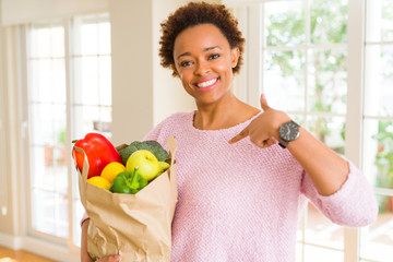 Young african american woman holding paper bag full of fresh groceries with surprise face pointing finger to himself