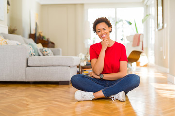 Young beautiful african american woman sitting on the floor at home looking confident at the camera with smile with crossed arms and hand raised on chin. Thinking positive.