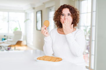 Senior woman eating healthy whole grain biscuit at home cover mouth with hand shocked with shame for mistake, expression of fear, scared in silence, secret concept